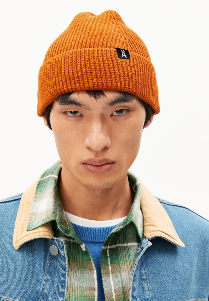 Orange knitted beanie with a textured ribbed pattern and a small black logo tag. Worn over a denim jacket with a plaid shirt collar.