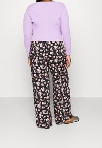 Woman wearing a lilac ribbed long-sleeve top, black wide-leg pants with pink floral print, and silver open-toe sandals, standing against a plain background.