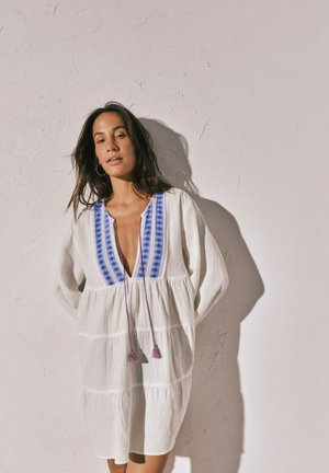 Frau mit langen dunklen Haaren, die ein weißes Kleid mit blau besticktem Ausschnitt und Quastenbändern trägt, steht vor einer strukturierten beigen Wand.