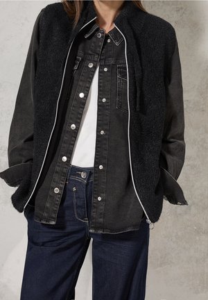 Person wearing layered clothing: black zip-up textured vest over black denim button-up shirt, white undershirt, and dark blue jeans with hands in pockets.