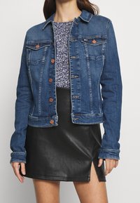 Jeanjacka med mörk tvätt, med två bröstfickor, kopparknappar, lång ärm och en kortare design. Bärs över en blommig topp.