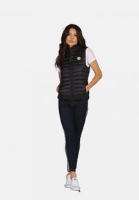 Gilet noir isolé avec un motif matelassé horizontal, col montant, fermeture éclair et patch de logo. Porté par-dessus un T-shirt blanc et associé à un jean sombre.