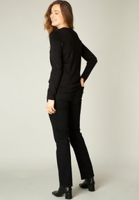 Zwarte aansluitende langemouwen top gecombineerd met zwarte rechttoe-rechtaan jeans. Model draagt zwarte haklaarzen en staat met de rug naar de camera met een neutrale achtergrond.