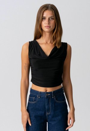 Tânără cu păr drept maro, purtând un top crop negru fără mâneci, drapat, și blugi albaștri cu talie înaltă, stând în picioare pe un fundal gri.