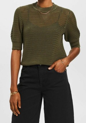 Femme portant un pull à manches courtes en maille ajourée vert olive, un pantalon noir taille haute, un collier chaîne en or et des bracelets, la main dans la poche.
