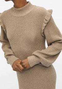 Gros plan sur une personne portant une robe en tricot beige avec des détails volants aux épaules et de longues manches bouffantes, les mains croisées à la taille.