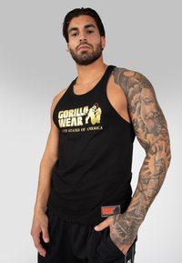Schwarzes ärmelloses Tanktop mit goldener "GORILLA WEAR"-Schrift und Logo. Verfügt über ein Marken-Patch und wird mit schwarzen Shorts getragen. Auffällige Armtattoos sind sichtbar.