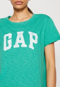 Grünes T-Shirt mit kurzen Ärmeln aus weichem Material, featuring ein großes weißes "GAP"-Logo mit einem leicht abgenutzten Look.