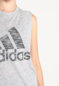 Harmaa hihaton paita, jossa on teksturoitu pinta ja iso musta Adidas-logo rinnassa. Sujuva kangas ja rento istuvuus.