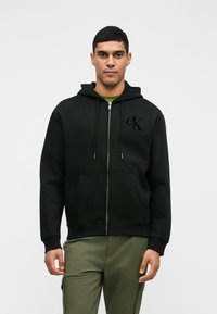 Μαύρο φούτερ με φερμουάρ και κορδόνια, με μαλακή υφή, διαθέτει ανάγλυφο λογότυπο "CK" στο αριστερό στήθος, συνδυασμένο με ελιά πράσινα παντελόνια.