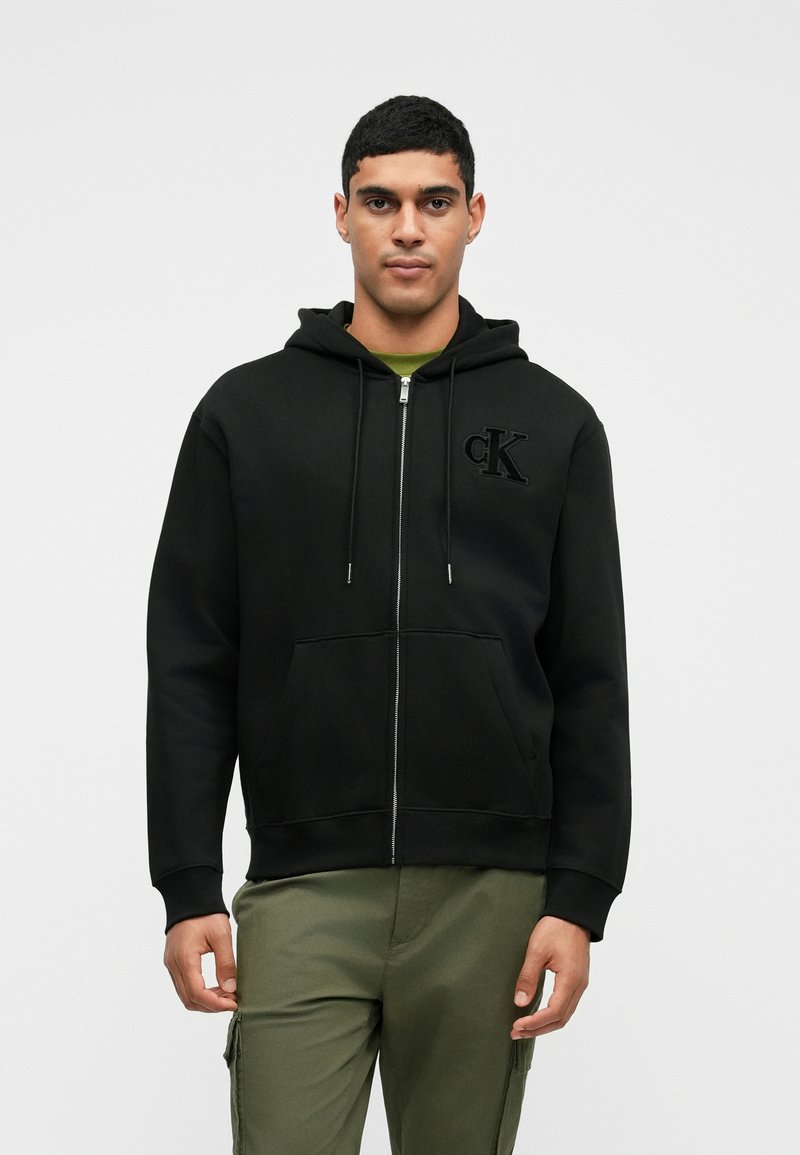 Μαύρο φούτερ με φερμουάρ και κορδόνια, με μαλακή υφή, διαθέτει ανάγλυφο λογότυπο "CK" στο αριστερό στήθος, συνδυασμένο με ελιά πράσινα παντελόνια.