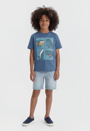 Laps lokkis juustega, kes kannab sinist T-särki, millel on neli multifilmilikku vaala ilustratsiooni, heledaid teksapükse ja tumesiniseid libisemisjalanõusid.