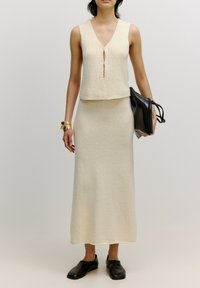 Beige gebreide twee-delige outfit met een mouwloze top met V-hals en een midi-rok. De stof heeft een gestructureerde afwerking. Zwarte clutchtas.