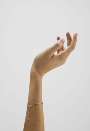 Mână dreaptă ridicată cu degete curbate, purtând o brățară delicată cu lanț auriu pe un fundal simplu gri deschis.