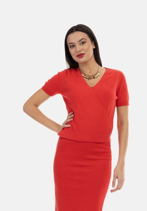 Rode gebreide wrap top met korte mouwen, V-hals en een getailleerd silhouet, gecombineerd met een bijpassende kokerrok. Gouden statement ketting.