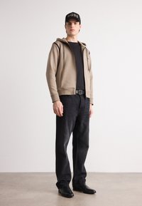 Haut à capuche beige zippé avec des accents noirs, porté sur un t-shirt noir. Un jean noir et des chaussures noires élégantes complètent la tenue. Casquette noire avec le logo "BOSS".