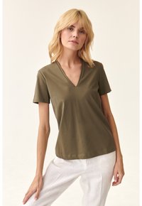 Blusa in cotone verde oliva con scollatura a V e maniche corte, caratterizzata da una vestibilità comoda e una texture liscia, abbinata a pantaloni in lino bianchi.