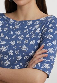 Blå blommönstrad topp med off-shoulder-design, tillverkad av mjukt tyg, med vita blommotiv och korta ärmar.
