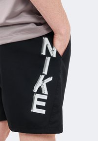 Άτομο που φοράει μαύρα σορτς με λευκό λογότυπο "NIKE" κάθετα, το χέρι ακουμπά στην δεξιά τσέπη, η κορμοστασιά με γκρι μπλουζάκι είναι ορατή.