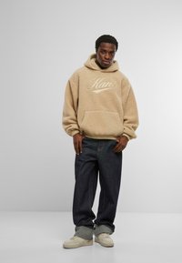 Beige polárkapucnis felső, dombornyomott logóval, elülső zsebbel és húzózsinórral. Sötét farmernadrággal és bézs tornacipővel párosítva.