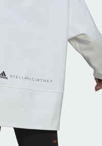 Vit långärmad topp med Adidas- och Stella McCartney-logotyper nära nederkanten, bärs med svarta byxor med detaljerade hål.