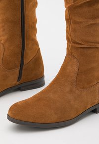 Bottes en suède marron montantes avec un design légèrement froissé, fermeture éclair sur le côté et bout arrondi. Talon plat avec une finition lisse.