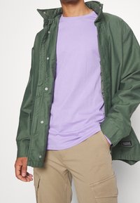 Groene waterdichte jas met drukknopen over een lavendelkleurig t-shirt. Beige cargobroek met zijzakken. Gladde stoftexturen overal.