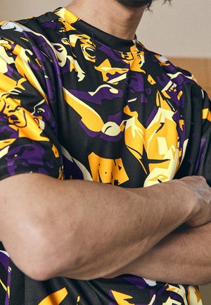 Persoon met gekruiste armen die een kleurrijk paars, geel en zwart grafisch T-shirt met abstracte patronen draagt.