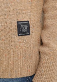 Beiger Strickpullover mit roten, blauen und gelben Sprenkeln. Mit geripptem Saum und Bündchen. Angesetztes schwarzes Etikett mit weißem Text und einem Gebäudegrafik.