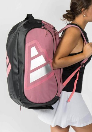 Vrouw draagt een zwart hemd en een witte rok en heeft een grote roze en zwarte sportrugzak met geometrisch patroon op haar rug.