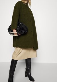 Cappotto verde oliva testurizzato con colletto alto e bottoni, abbinato a una gonna in satin beige e stivali neri al ginocchio, con una borsa nera arricciata in mano.