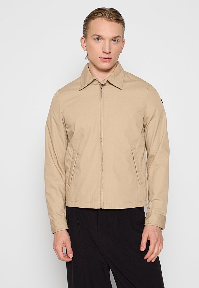 Veste légère beige avec fermeture éclair à l'avant, col pointu et poches latérales. Présente une texture lisse et un accent de logo noir sur la manche.