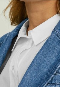 Džínová bunda s modrým omytím, s špičatým límcem a světle barevnou košilí na knoflíky pod ní. Texturovaná látka, viditelné kontrastní švy.