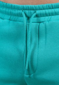 Turkusowe spodnie dresowe z elastycznym pasem, regulowanym sznurkiem i gładką, bawełnianą tkaniną. Posiadają boczne kieszenie.
