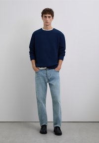 Granatowy sweter z dzianiny z fakturowanym wzorem, jasno-niebieskie dżinsy o prostych nogawkach oraz czarne buty. Minimalistyczny design na neutralnym tle.