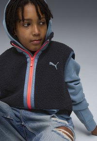Dunkelblaues Fleece-Weste mit hellrosa Reißverschluss und hellblauer Kapuzen-Basislagenschicht. Abgenutzte Jeans aus Denim. Puma-Logo auf der Weste.