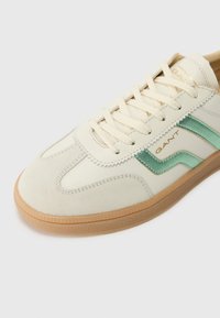 Hvit og tan sneakers med overdel i semsket skinn og lær, med en grønn stripe og strukturert gummisåle. Snøringene er lys beige.