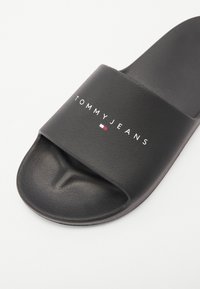 Crna klizna sandala napravljena od teksturiranog sintetičkog materijala, sa širokom trakom na kojoj je bijeli natpis "TOMMY JEANS" i crveno-bijelim logotipom.