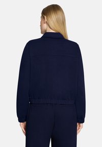 Granatowa bluza z teksturą jednolitą, kołnierzem, elastycznym pasem i długimi rękawami. Charakteryzuje się prostym designem z minimalną ilością detali.