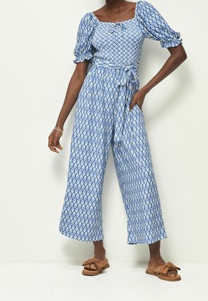 Femme en combinaison bleue et blanche à motifs avec manches bouffantes, jambes larges et ceinture nouée à la taille, portant des sandales marron à enfiler.
