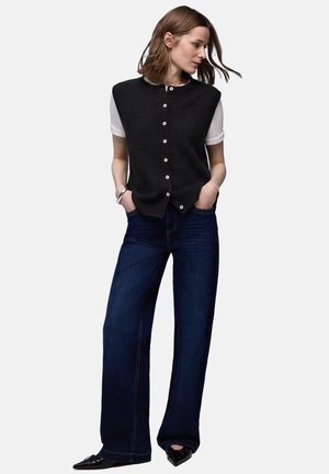 Sort strikket ærmeløs top med knaplukning, båret over en hvid skjorte. Kombineret med mørkeblå vidde-bukser og sorte spidse sko.