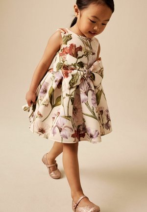 Ung pige iført en ærmeløs blomstret kjole med en stor sløjfe, gående i glitrende lyserøde sko på en neutral baggrund.