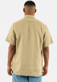 Camicia khaki a maniche corte, tessuto leggero, taglio dritto, dettaglio sulla parte posteriore, aperture laterali per facilitare i movimenti, texture morbida.