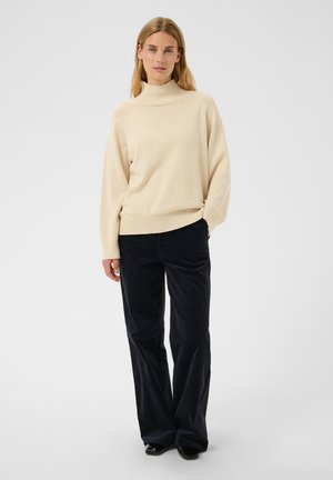 Femme portant un pull à col roulé beige et un pantalon noir à jambes larges, debout devant un fond blanc uni.