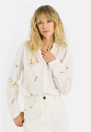 Mujer rubia con cabello ondulado, vestida con blusa blanca bordada y pantalones blancos, con la mano apoyada en el pecho, fondo neutro.