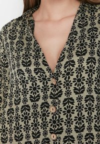 Svart blommönstrad blus på olivgrön tyg, med V-hals och träknappar, som framhäver dess intrikata design och textur.