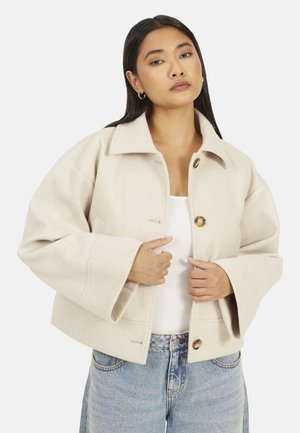 Femme aux cheveux raides et foncés portant des boucles d'oreilles anneaux dorées, une veste en laine beige boutonnée, un haut blanc et un jean bleu clair.