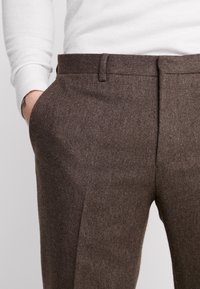 Man in bruine wollen broek met hand in de linker broekzak en een wit langemouwen shirt zichtbaar van taille tot pols.