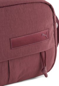Borsa in tessuto color mogano con una superficie liscia, caratterizzata da un logo Puma ben visibile, una tasca esterna e una chiusura con coulisse per maggiore comodità.