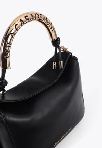 La borsa nera presenta un manico dorato prominente inciso con "LOLA CASADAMUNT" e un corpo in pelle liscia, evidenziando un'eleganza moderna.